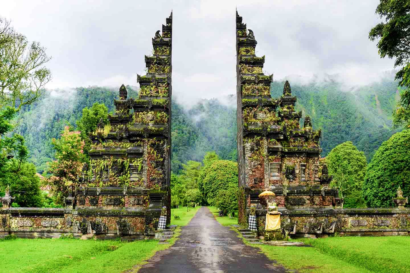 Ancient stone temple gates with ornate carved details covered in moss, surrounded by lush green rice fields and misty mountai